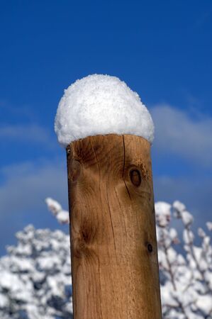 Freshly fallen snow is piled up about five inches on top of a wooden fence pole.  Blue sky and snowy branches form background.の写真素材