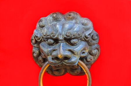 Detailed brass door knocker has lion's head in a Chinese design.  Ring hangs from its mouth and it is mounted on a red door.の写真素材