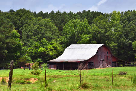 Red faded barn has tin roof.  Exterior has broken boards.  Farm and pasture is surrounded by a...