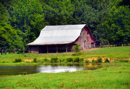 Large, red wooden, barn sits in green pasture with a pond.  Boards are faded and broken.