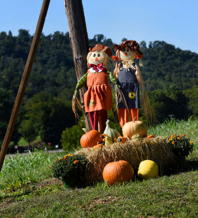 Two scarecrows stand behind a bale of hay, pumpkins and squash.  Fall decorations spruce up a country farm.の写真素材