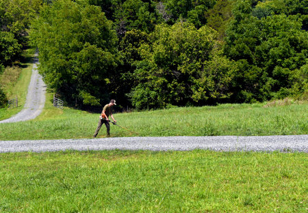 Young man works to trim gravel driveway.  It is Summer and grass is green.  Driveway descends hill...