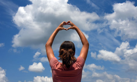 Young, female, teen forms heart shape with her hands.  She is looking into a blue sky withbig fluffy...