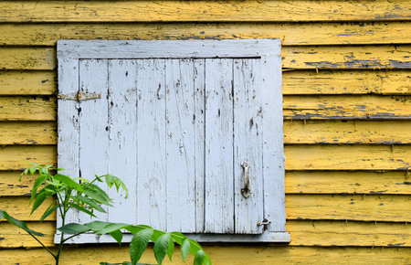 Rustic white, wooden, window is shuttered and hooked.  Window is on a bright yellow building with peeling paint.の写真素材