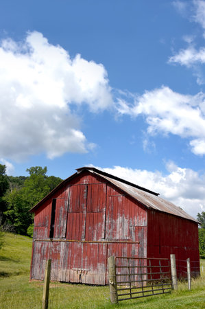 Weathered and faded, wooden, red barn sits besides a wooden fence row.  Farm is in Tennessee.  Blue skies overhead.の写真素材