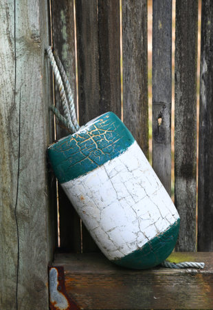 Fishing buoy leans against a rustic wooden fence. It's green and white striped surface has been weathered by the sun and ocean water.の写真素材