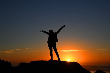 Excited woman reaches her arms wide to embrace a glorious, golden sunset on Bullards Beach State Park.  She is standing on a rocky bluff besides the beach.の写真素材