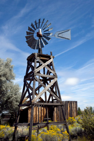 Rustic, wooden windmill stands against a blue sky.  Yellow Sage and Rabbits Brush cover wooden legs.