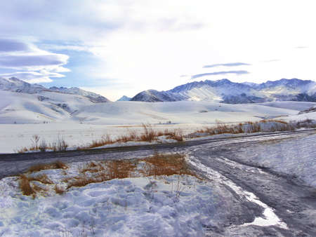 Snowy mountains and ridge, country road with puddle, brown grass in the foreground. Winter landscape. Copy spaceの写真素材