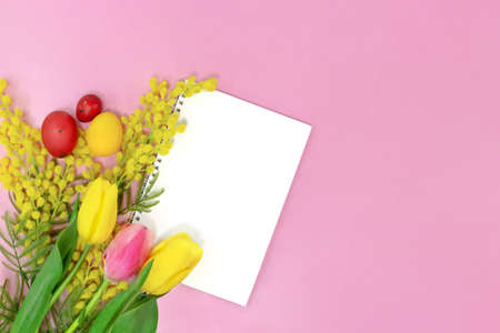 Bouquet of flowers, tulips and yellow mimosa with blank sheet of paper for text on pink background with colored eggs. Easter background. Copy spaceの写真素材