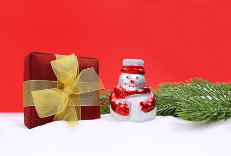 Snowman with present box in snow and green spruce branches on red background. Christmas, New Year. Copy spaceの写真素材
