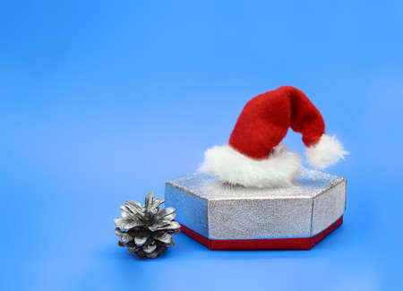 Silver gift box with Santa Claus hat and pine cone on blue background. Christmas, New Year. Copy spaceの写真素材