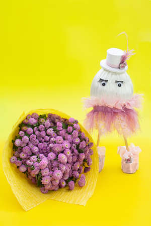 Bouquet of lilac chrysanthemums and girl egg in skirt and hat on yellow background. Easter, birthday, March 8, International Women's, mother's day. vertical. copy spaceの写真素材