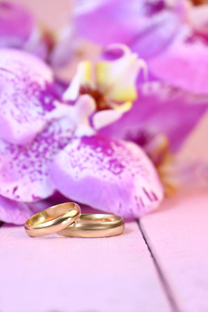 Golden wedding rings with purple orchids on pink wooden table. vertical. Wedding ceremony, honeymoon, marriage proposal, Valentine's day in hot countries. copy spaceの写真素材