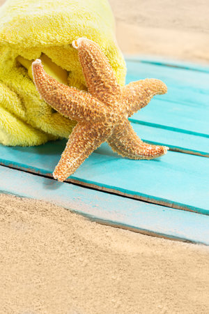 Orange starfish with yellow towel on blue wooden board of pier or sun lounger on sand of beach. Vacation, travel, weekend. copy spaceの写真素材