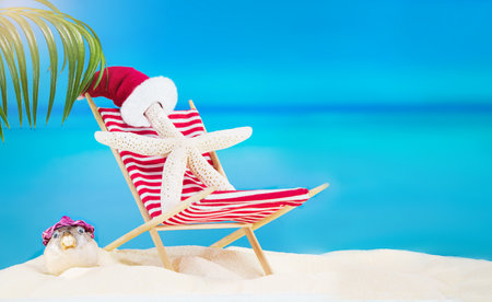 White starfish with Santa' hat on striped red sun lounger with puffer fish on sandy beach with palm tree behind sea. Christmas, New Year, summer, travel, vacation, holiday in hot countries. Copy spaceの写真素材