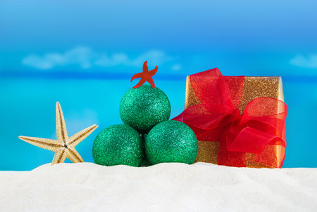Green shiny Christmas tree made of glitter balls with starfish, wrapped gift box with red bow on sand of beach, sea behind. New Year, holidays in hot countriesの写真素材