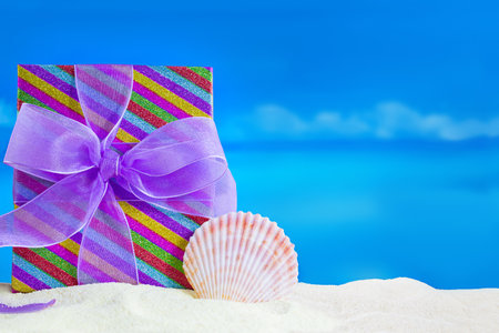 Striped gift box with bow and conch on sandy beach, behind sea. Vacations, relaxation, travel to hot countries. Christmas, New Year, birthday, Mother's and Women's Day, congratulations, holiday. Copy spaceの写真素材