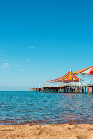 Wooden pier with orange-red awning over water of sea, lake, ocean with beach without people. Travel, vacation, summer background. Vertical. Copy spaceの写真素材