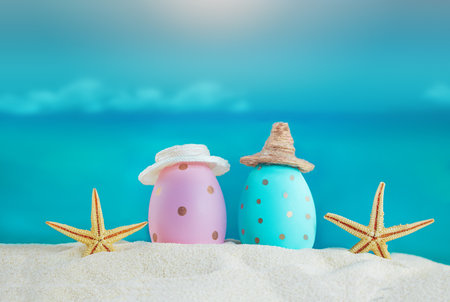 Blue and pink eggs in hats with starfish on the sand of the beach, beyond the sea. Easter in hot countries. Copy spaceの写真素材
