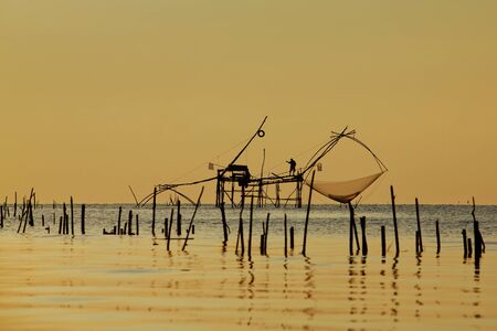 Asian fisherman on wooden boat casting a net for catching freshwater fish in nature river in the early morning with sunriseの写真素材