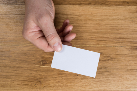 Hand holding white blank paper sheet mockup on wooden background. Arm in shirt hold clear brochure template mock up. Leaflet document surface design. Simple pure print display show. Reading contract agreement.の写真素材