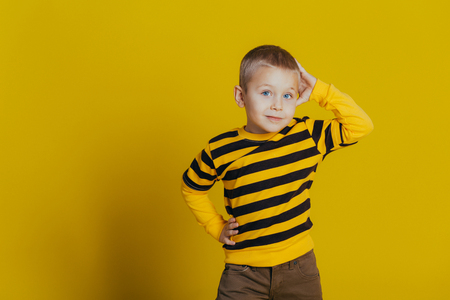 Portrait of an attractive thoughtful boy in a striped sweater holding a hand on his head close-up, on a yellow backgroundの写真素材