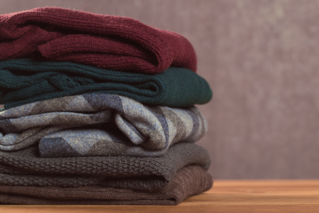 Bunch of knitted warm pastel color sweaters with different knitting patterns folded in stack on brown wooden table, grunged texture wall backgroundの写真素材
