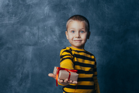 A young emotional boy holds in his hands a gift with a red bow standing on a blue studio...
