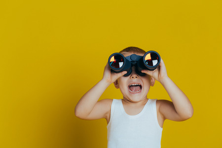 Portrait of an attractive smiling boy in a white vest, looking through black binoculars close-up, on a yellow backgroundの写真素材