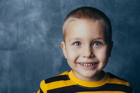 Little fashionable boy posing in front of a gray-blue concrete wall. Portrait of a smiling child dressed in a black and yellow striped sweater and brown pants.の写真素材