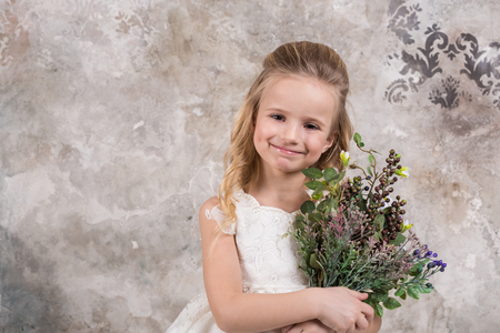 Portrait of a little attractive girl in a white dress with a bouquet in her hands against the background of a grunge wall. Studio shot with space for text.の写真素材
