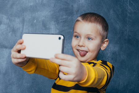 little cute boy with a mobile phone takes a selfie and shows emotions on a blue backgroundの写真素材