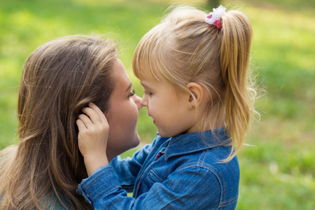 little happy girl hugs her mom and tells her something in the ear in the parkの写真素材