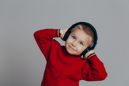 Portrait of an attractive smiling boy in a red sweater listening to music through headphones close-up, on a gray backgroundの写真素材