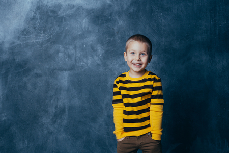 Little fashionable boy posing in front of a gray-blue concrete wall. Portrait of a smiling child dressed in a black and yellow striped sweater and brown pants.の写真素材
