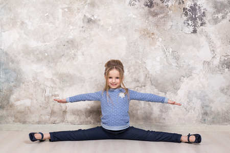 Portrait of a little attractive smiling girl in a blue sweater and pants with her hair folded in her hair sitting on a string against the background of a grunge wall. Studio shot with place for text, fashion conceptの写真素材