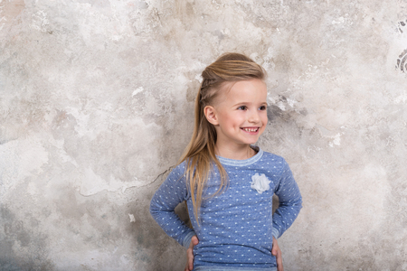 Portrait of a little attractive smiling girl in a blue sweater and pants with hair folded into her hair against a grunge wall background. Studio shot with place for text, fashion conceptの写真素材