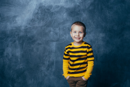Little fashionable boy posing in front of a gray-blue concrete wall. Portrait of a smiling child dressed in a black and yellow striped sweater and brown pants.の写真素材