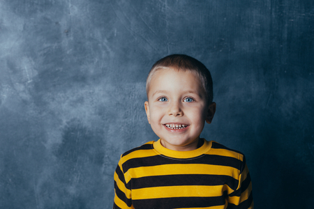 Little fashionable boy posing in front of a gray-blue concrete wall. Portrait of a smiling child dressed in a black and yellow striped sweater and brown pants.の写真素材