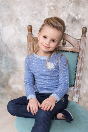 Portrait of a little attractive smiling girl in a blue sweater and pants with her hair folded in her hair sitting on a chair against a grunge wall background. Studio shot with place for text, fashion conceptの写真素材