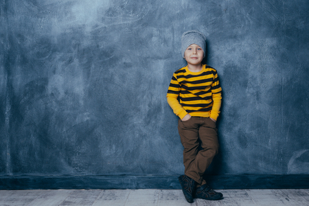 Little fashionable boy posing in front of a gray-blue concrete wall. Portrait of a smiling child in a black and yellow striped sweater and brown trousers. The concept of style and fashion for children.の写真素材