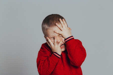 Portrait of an attractive boy in a red sweater holding his head with his hands close-up, on a gray backgroundの写真素材