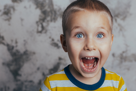 A little boy poses in front of a gray concrete wall. Portrait of a screaming child with open mouth, dressed in a black and yellow striped jacketの写真素材
