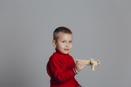 Portrait of attractive boy in red sweater playing with wooden plane close-up, on gray backgroundの写真素材