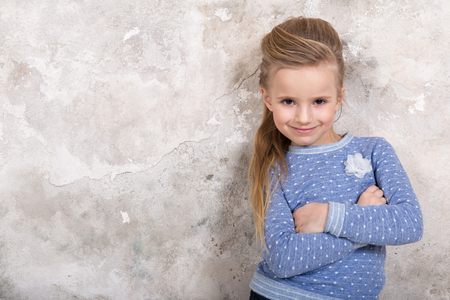 Portrait of a little attractive smiling girl in a blue sweater and pants with hair folded into her hair against a grunge wall background. Studio shot with place for text, fashion conceptの写真素材