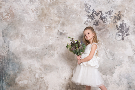 Portrait of a little attractive girl in a white dress with a bouquet in her hands against the background of a grunge wall. Studio shot with space for text.の写真素材