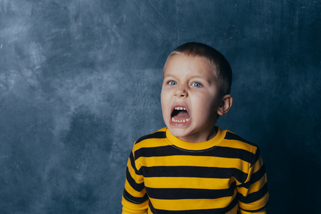 A little boy poses in front of a gray-blue concrete wall. Portrait of a screaming child with open mouth, dressed in a black and yellow striped jacketの写真素材