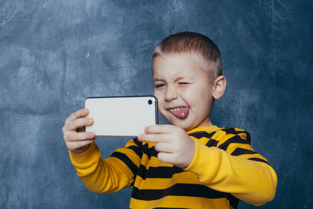 little cute boy with a mobile phone takes a selfie and shows emotions on a blue backgroundの写真素材