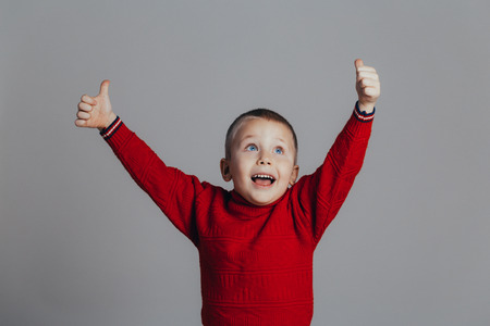 Portrait of an attractive boy in a red sweater who raised his hands up close-up, on a gray backgroundの写真素材